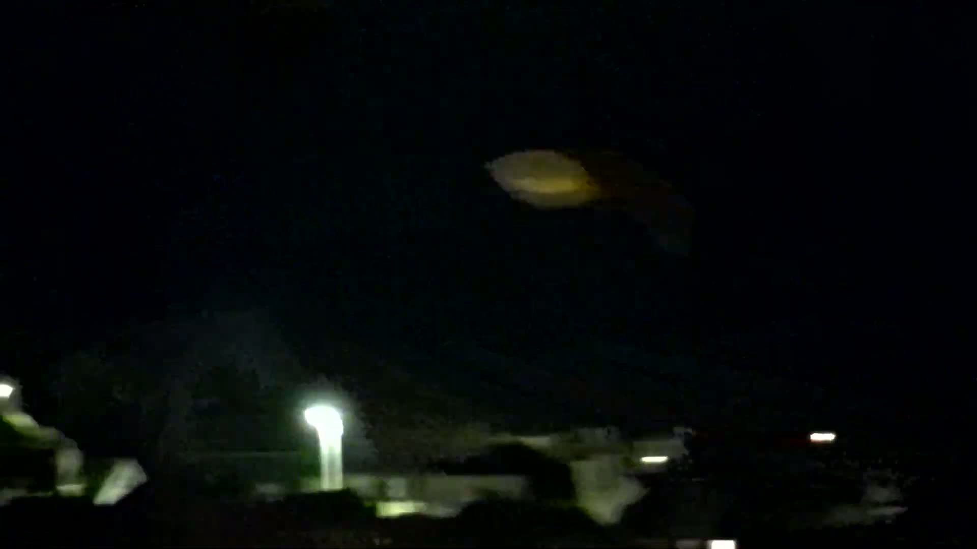 A blurry, yellowish-orange object is visible in the night sky above a dimly lit residential area.