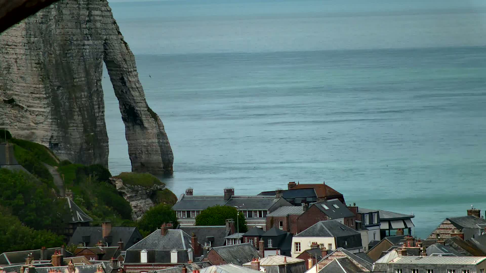 A large, grey, arch-shaped cliff overlooks a village of dark-roofed houses beside a calm, blue-green ocean under an overcast sky.