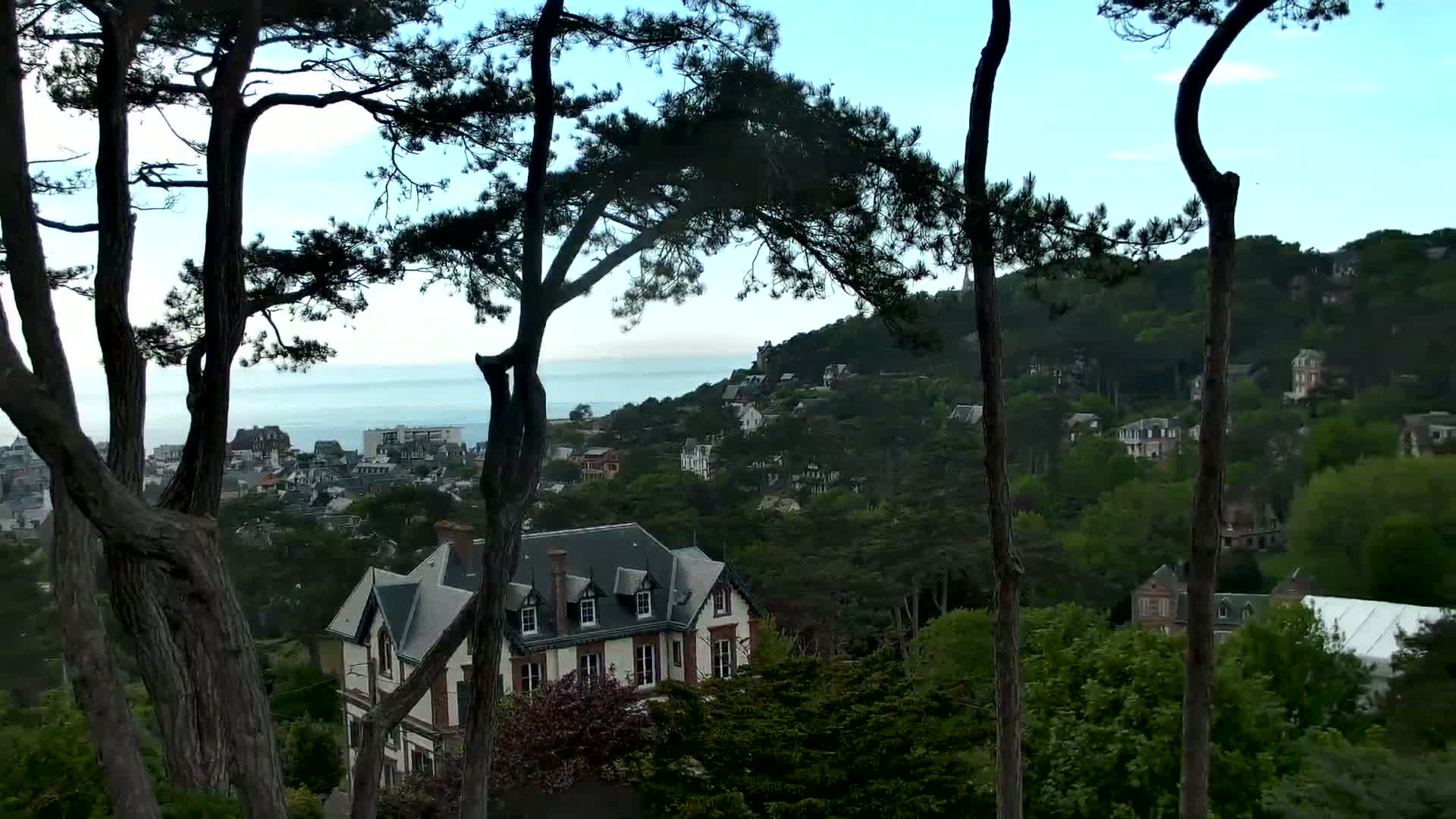 A large white and red house sits nestled among a hillside of lush green trees, overlooking a town and calm ocean under a clear sky.