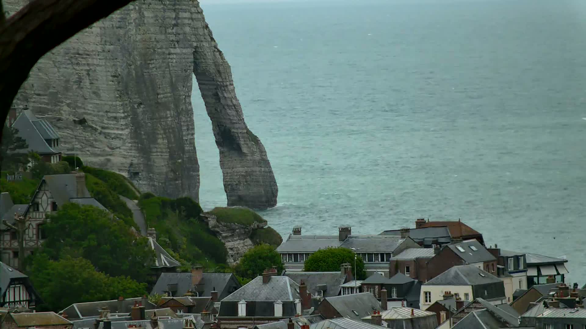 A large, gray, elephant-shaped cliff overlooks a village of dark-roofed houses by a choppy, gray-green sea under an overcast sky.