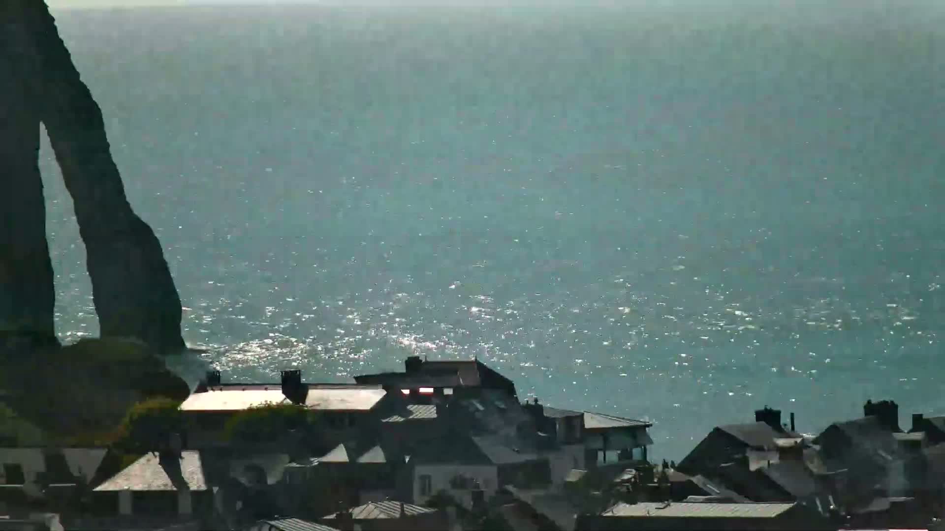 A cluster of dark-roofed houses sits atop a cliff overlooking a sparkling, teal ocean under a bright, hazy sky.