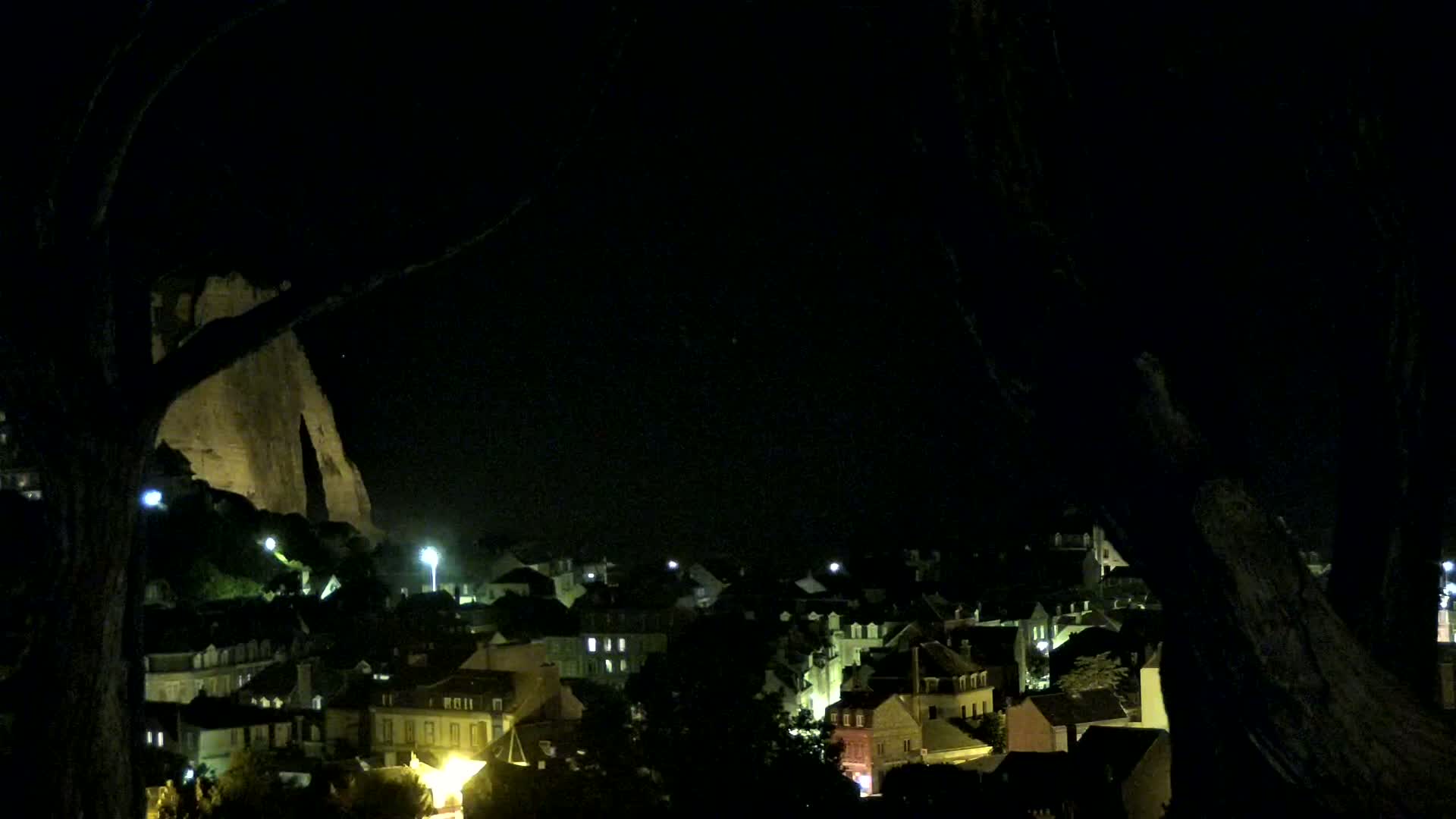 At night, a town's illuminated buildings are visible beneath a large rock formation, partially obscured by silhouetted trees.