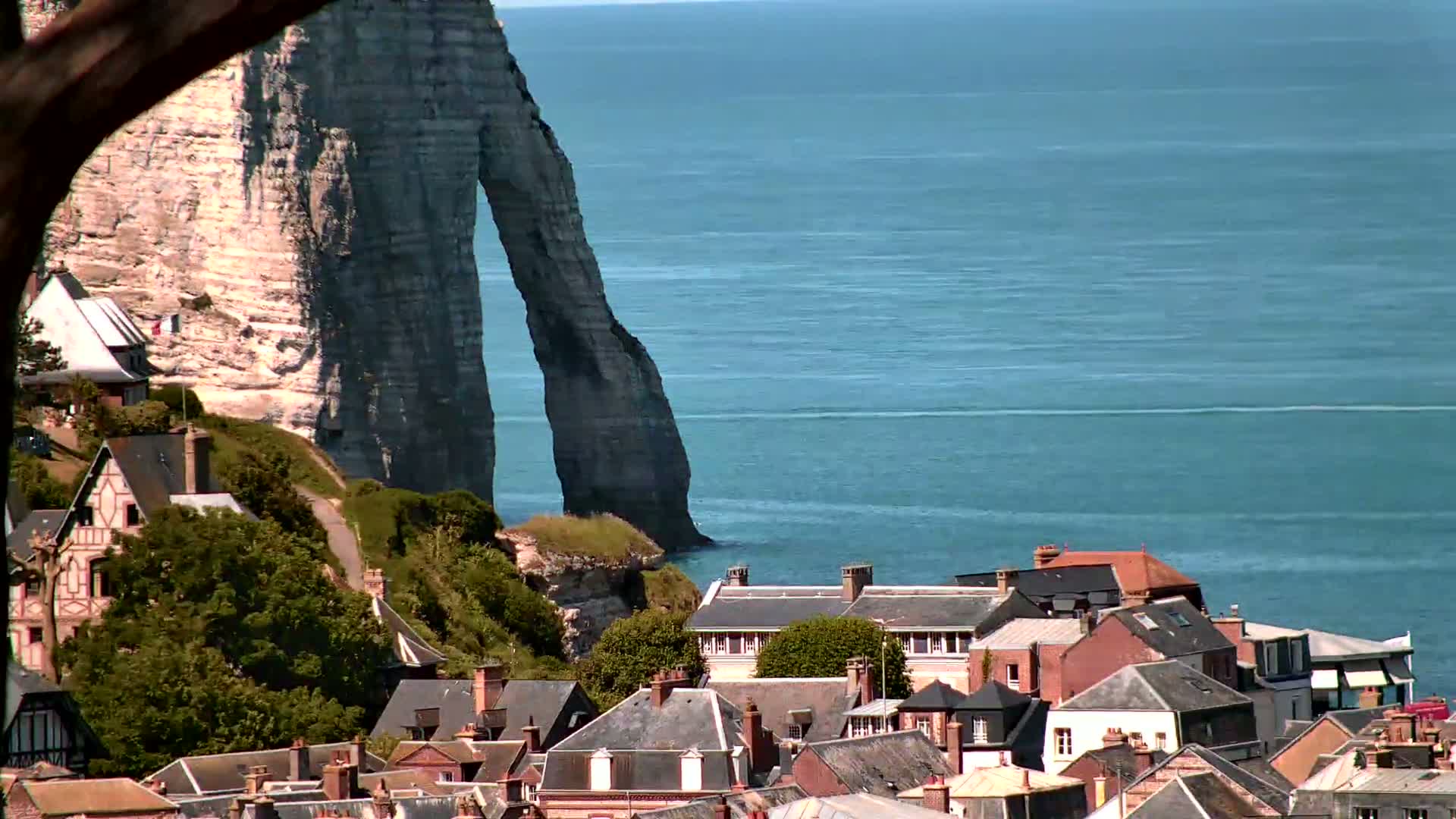 A coastal village with houses of varying architectural styles nestles beneath a dramatic, naturally arched white cliff overlooking a calm blue sea on a sunny day.
