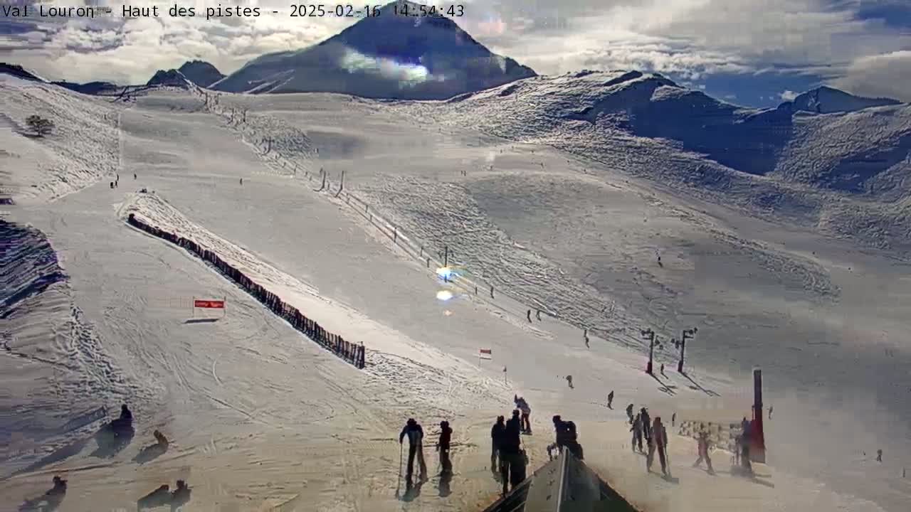 A snowy mountain slope with several skiers and snowboarders visible on the piste under a partly cloudy sky.
