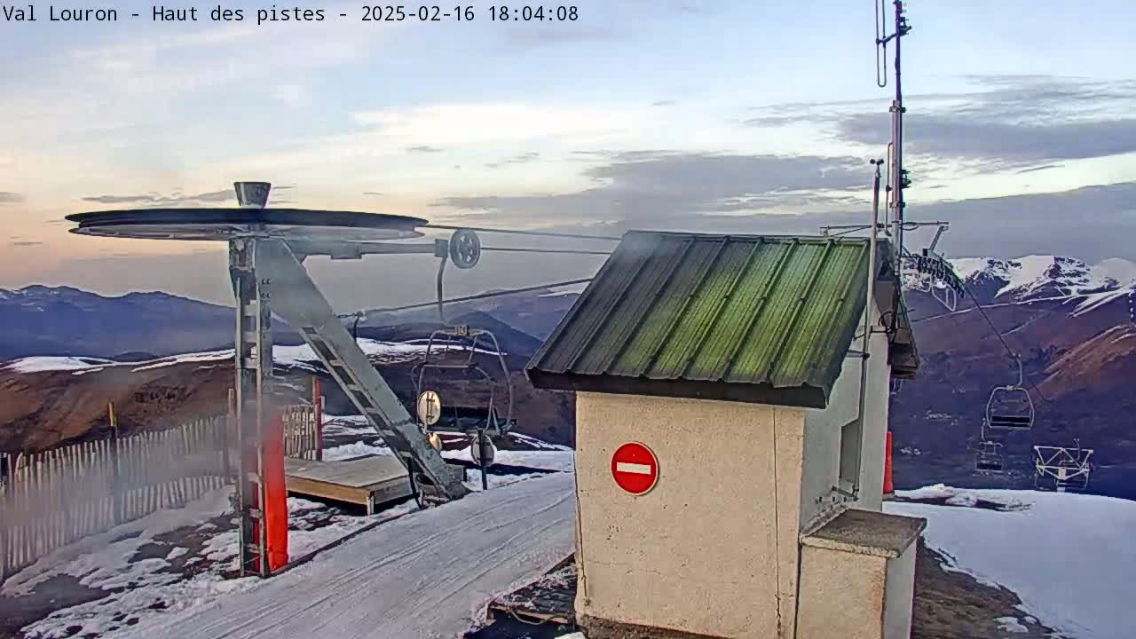 A ski lift's pulley and control station are situated on a snowy mountaintop under a partly cloudy sky, with empty ski lift chairs visible in the distance.