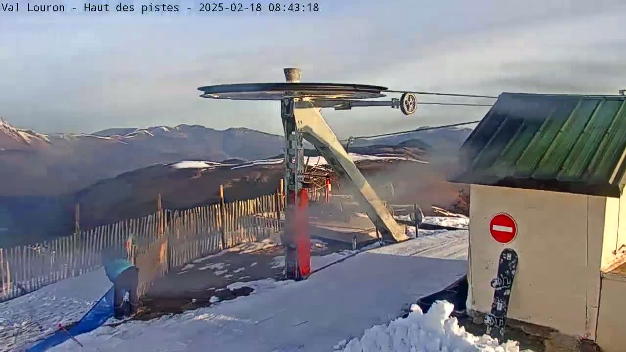 A ski lift pulley system is seen on a snowy mountaintop with a partly cloudy sky and a person standing nearby.