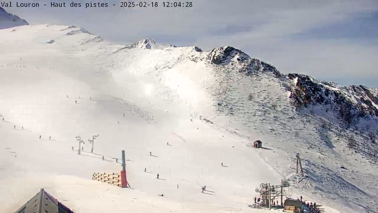 A snow-covered mountain slope with skiers and snowboarders scattered across the piste under a partly cloudy sky.