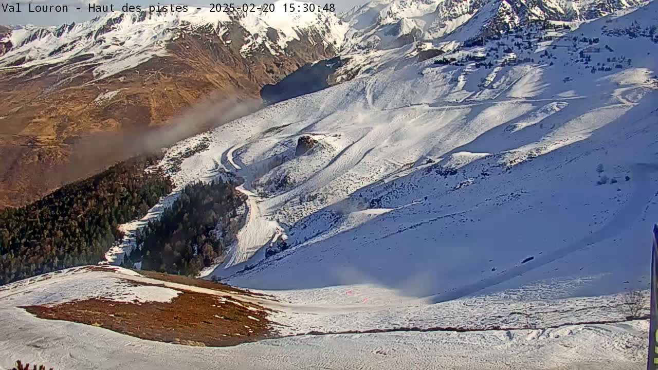 A high-angle view shows a snow-covered mountain slope with ski runs, a dark green forest below, and a hazy, brown valley in the background under a sunny sky.