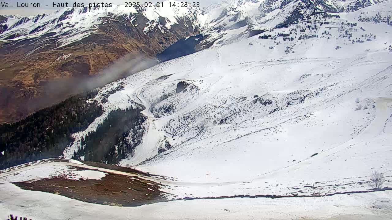 A snow-covered mountain slope with some patches of dark earth and trees, viewed from above, with a light haze or fog in the distance.
