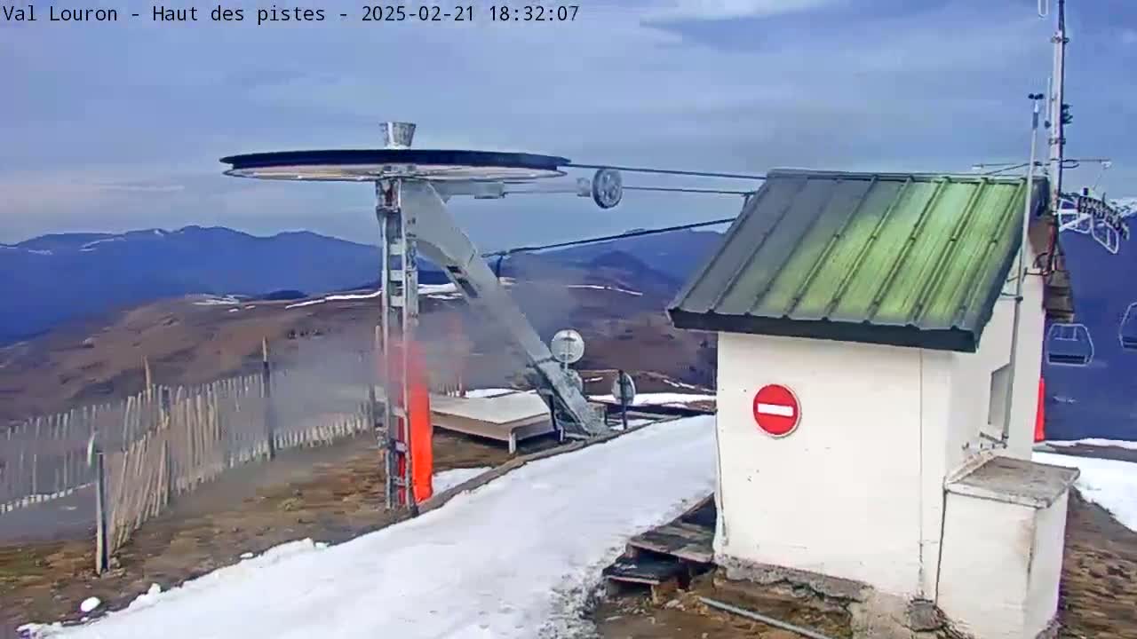 A ski lift mechanism and a small building are situated on a snow-covered mountaintop under a mostly clear sky.