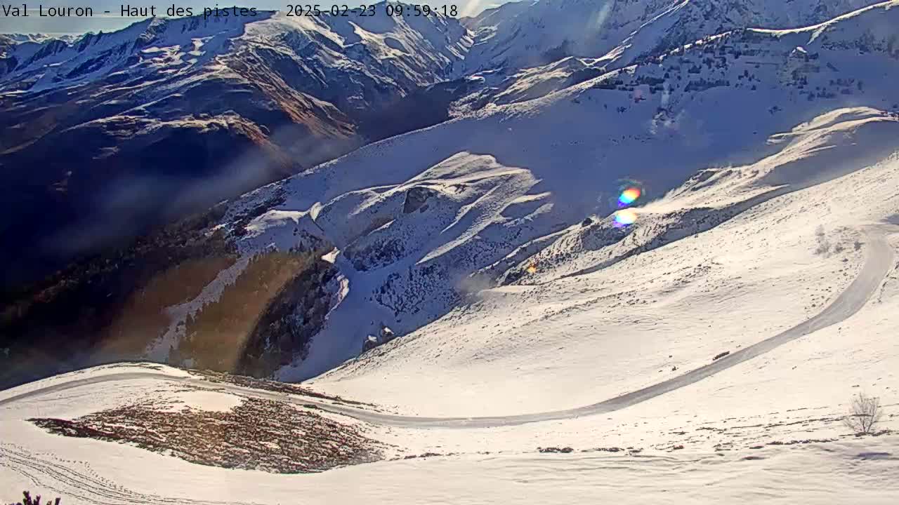 A winding snow-covered road snakes through a sunny, snow-capped mountain range.