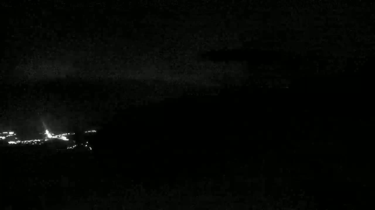 A low-light nighttime image shows a collection of faint lights in the distance, possibly a town or city, under a dark, cloudy sky.
