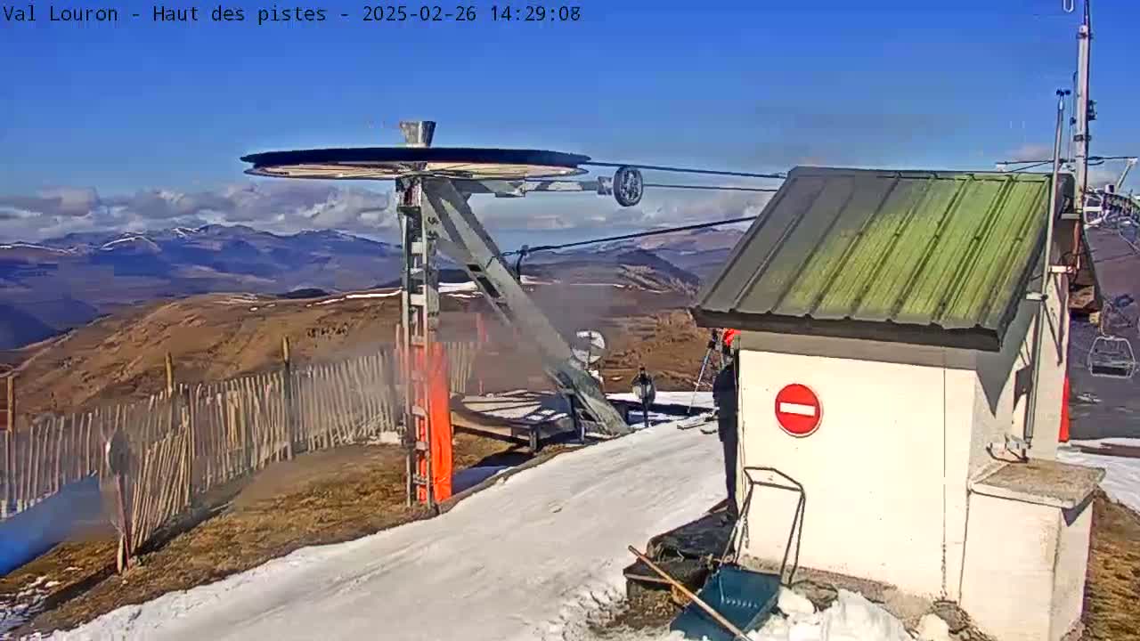 A ski lift pulley system and small building are situated on a snowy mountain slope under a mostly clear, sunny sky with distant mountains visible in the background.