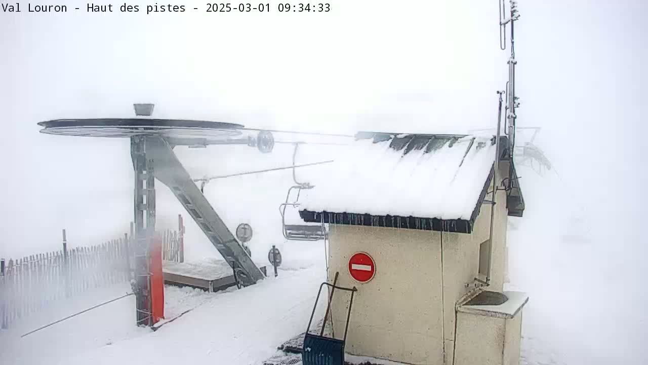 A snow-covered ski lift station is shrouded in fog, with a portion of the lift mechanism and a snow-covered building visible.