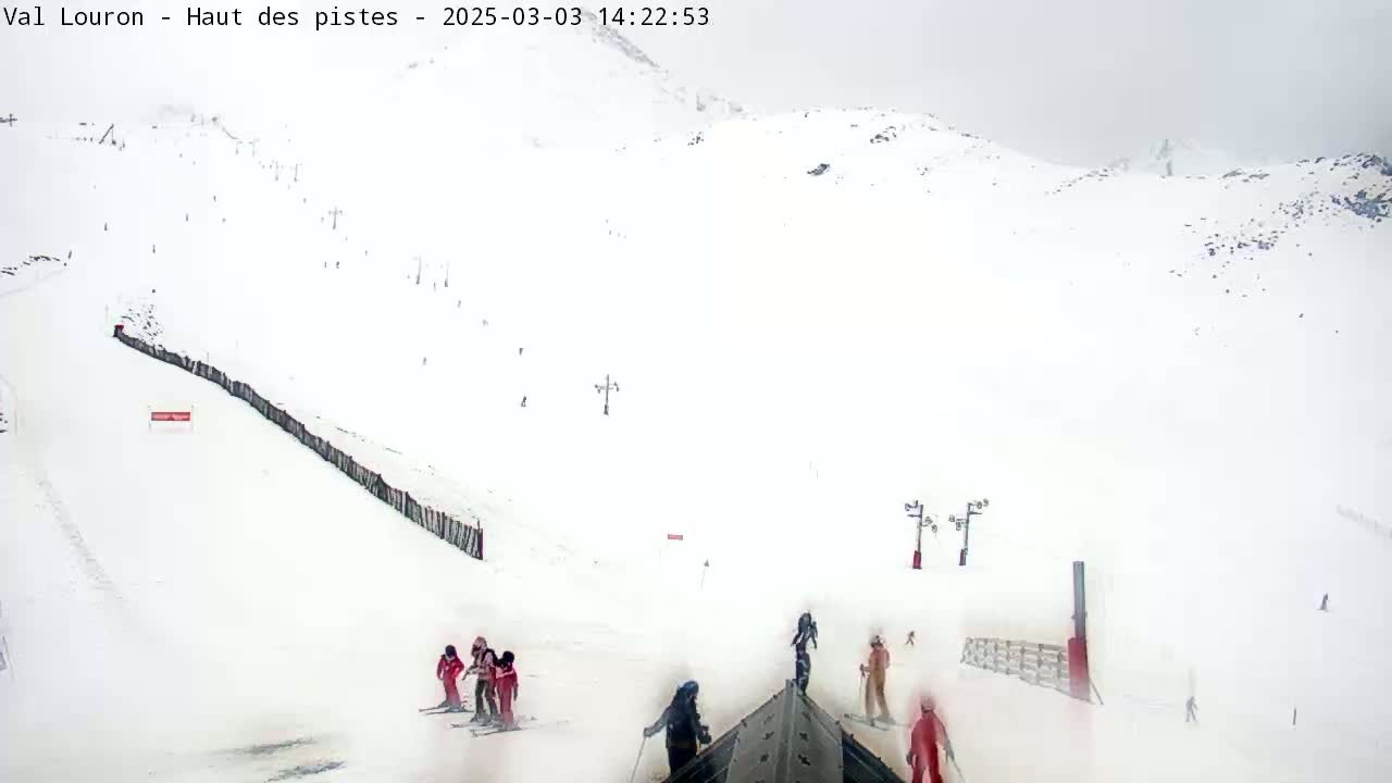 A snowy, overcast ski slope with several skiers and snowboarders visible, along with ski lifts and fences.