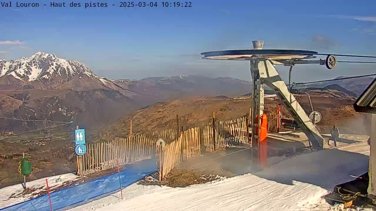 A ski lift sits atop a snow-covered mountain with a clear, sunny sky and a snow-capped mountain range in the background.