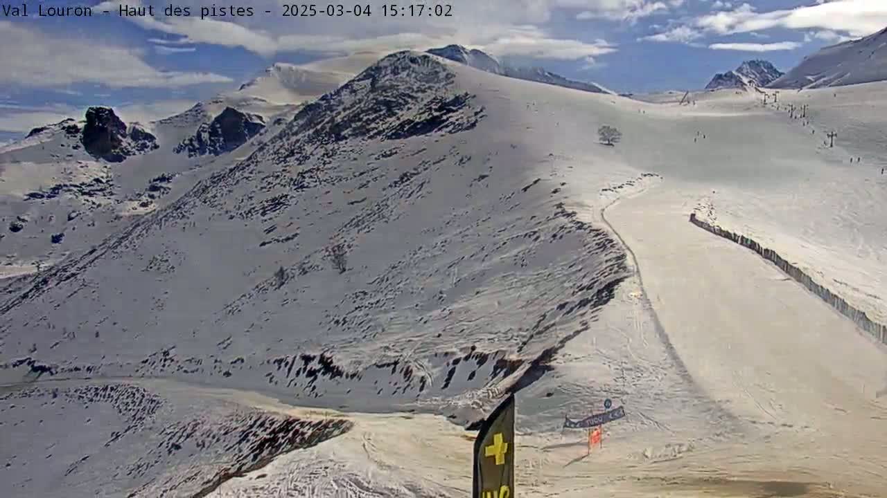 A snow-covered mountain slope with a ski run, under a partly cloudy sky.