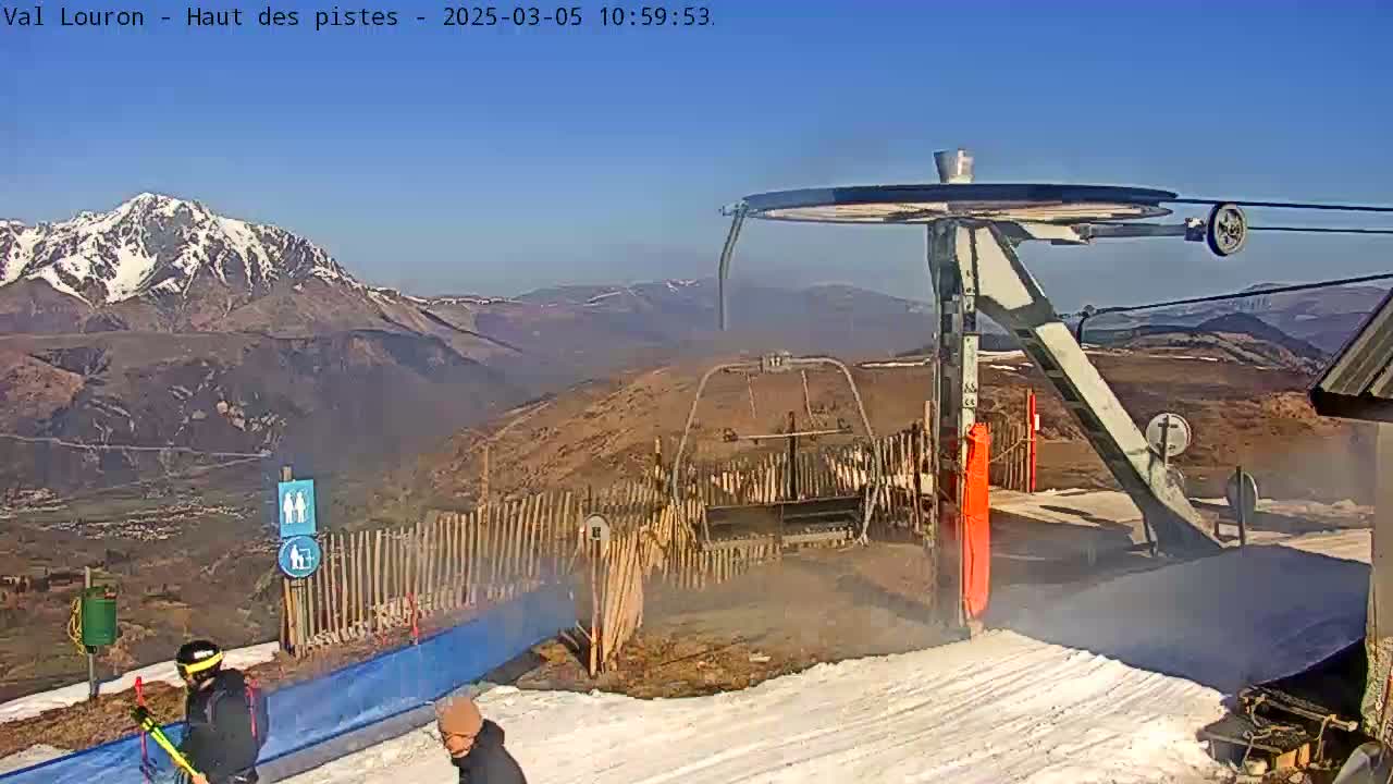 A ski lift base station with two people nearby, overlooking a snow-capped mountain range on a clear, sunny day.