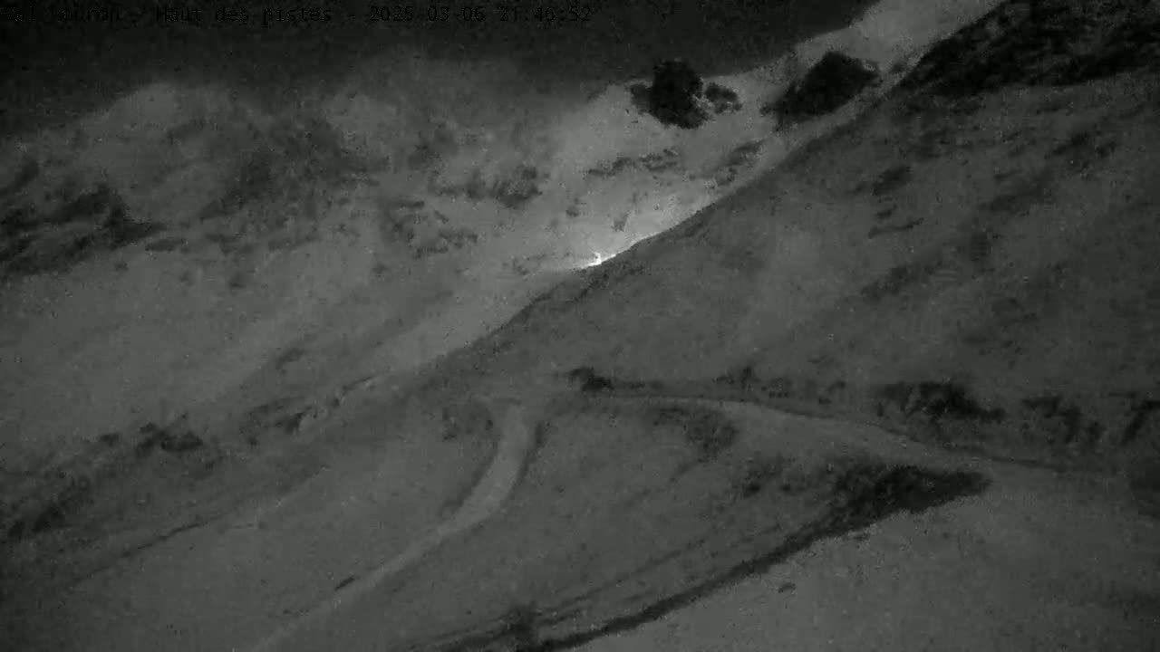 A nighttime, snowy mountain road winds through a snow-covered landscape under a cloudy sky, with a faint light visible in the distance.