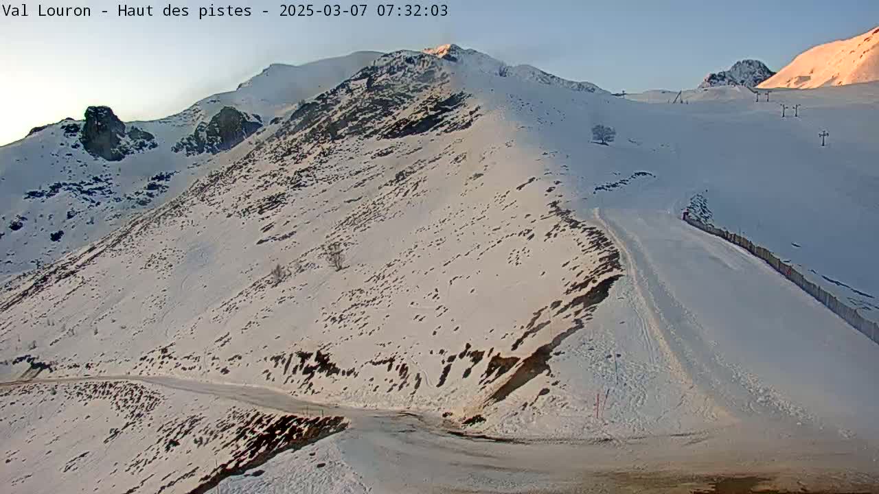 A snow-covered mountain slope with a ski run and a road winding down, under a clear, bright sky at sunrise.