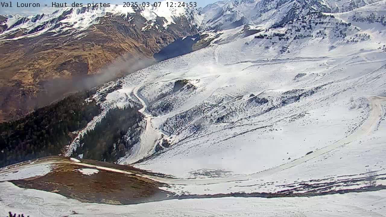 A snow-covered mountain slope with a winding road and patches of dark earth is visible, under a mostly clear sky.