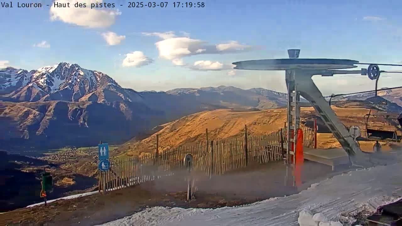 A ski lift stands atop a snow-dusted mountain range under a partly cloudy sky.