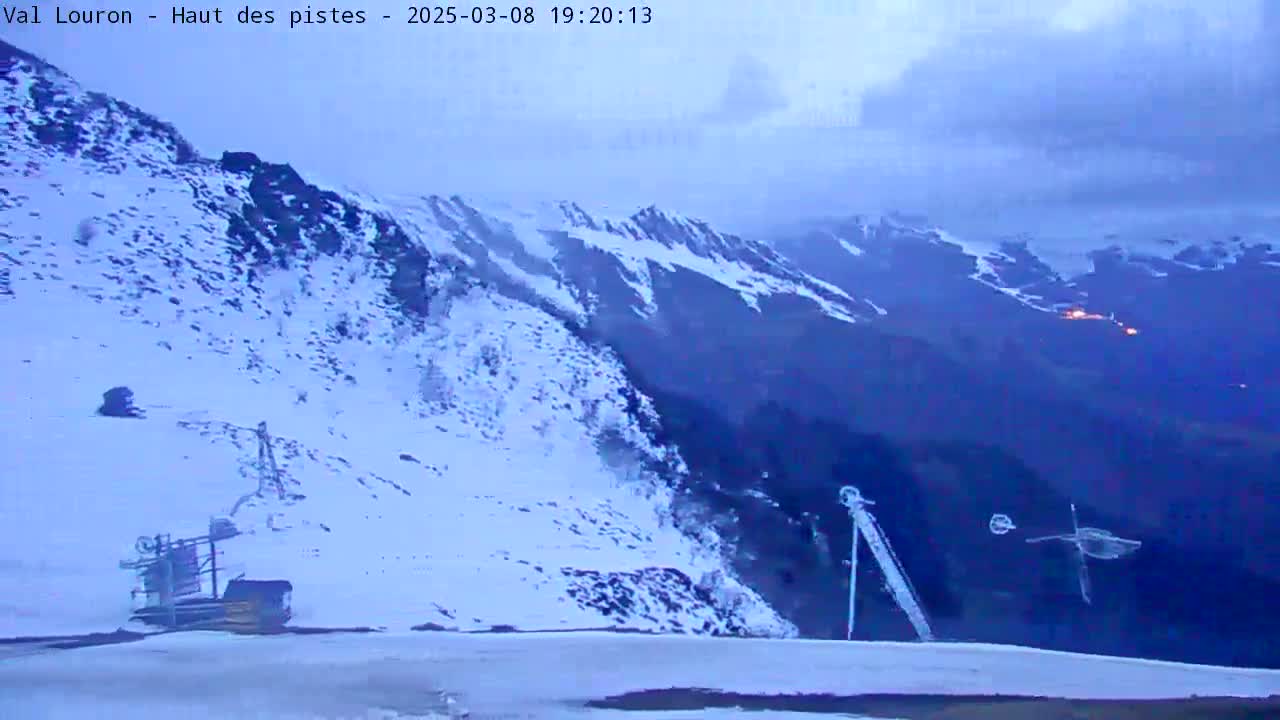 A snow-covered mountain slope at dusk, with a ski lift in the foreground and distant mountains under a cloudy sky.