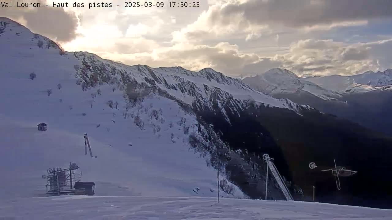 A snow-covered mountain slope with ski lift equipment under a partly cloudy sky.