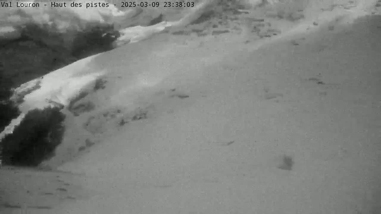 A nighttime, grayscale image shows a snow-covered slope with a darker area at the bottom, possibly a wooded area.