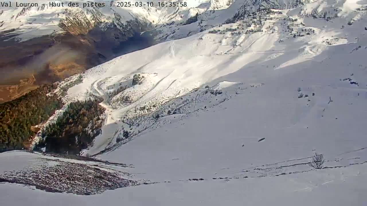 A snow-covered mountain slope with patches of dark-colored vegetation and a winding road or trail visible in the mid-ground, under a bright, sunny sky.