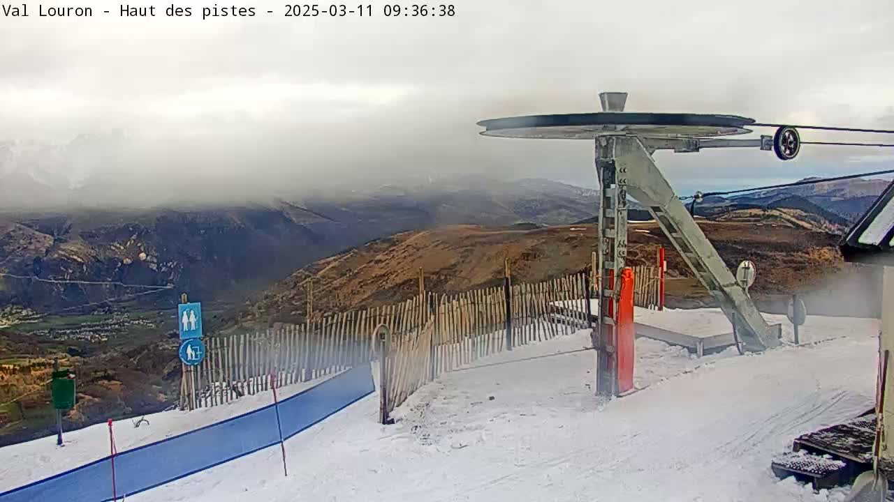 A ski lift pulley system is situated on a snow-covered mountaintop overlooking a valley under an overcast sky.
