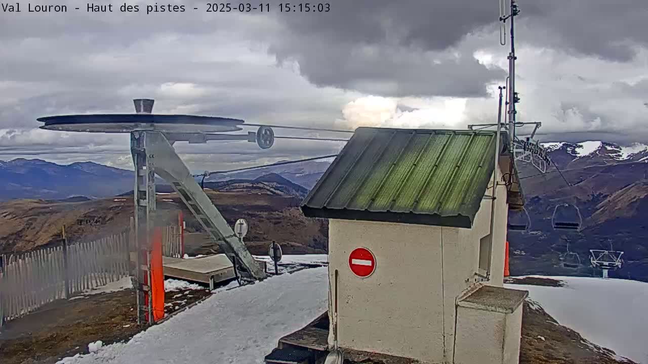 A ski lift pulley system and small building are situated on a snowy mountaintop under a cloudy sky, with distant mountains visible in the background.