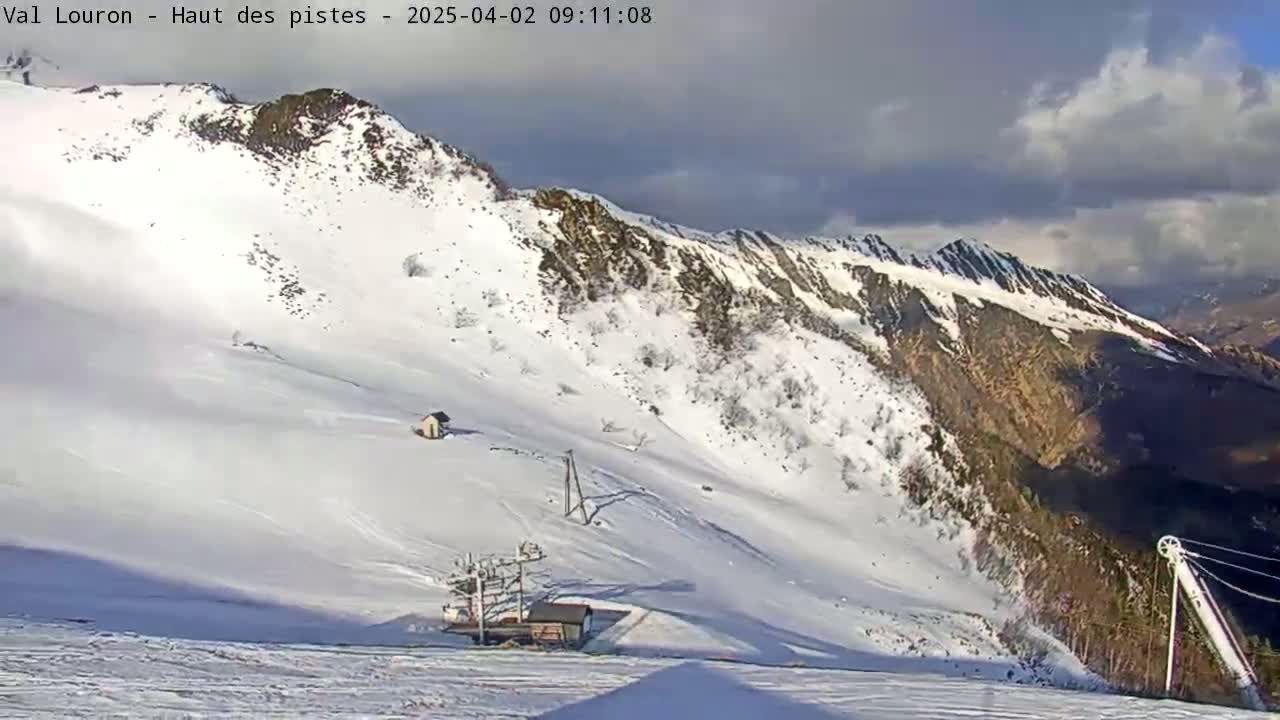 A snow-covered mountain slope with a partly cloudy sky shows a ski lift and small buildings under partly sunny conditions.