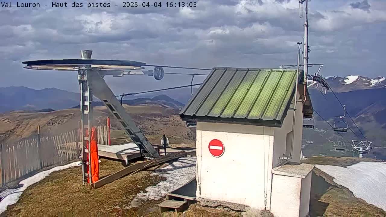 A ski lift's top station, under a partly cloudy sky, overlooks snow-dusted mountains in the distance.