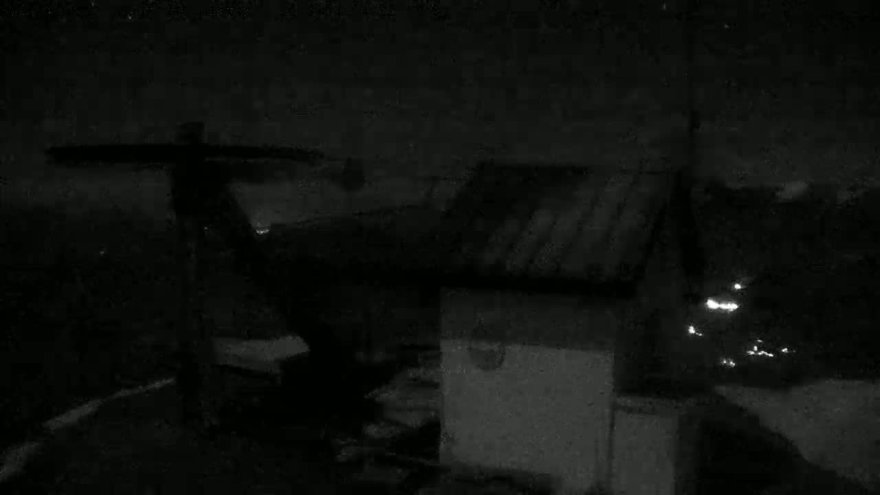 A low-light nighttime image shows a dark structure with a sloped roof and a large, possibly rotating, object attached to its side, under a dark, cloudy sky.