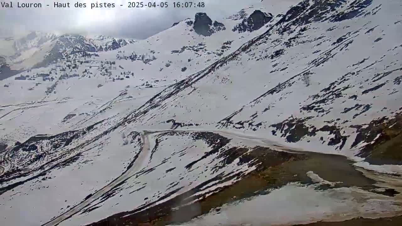 A snow-covered mountain range with a winding road visible, under overcast skies.