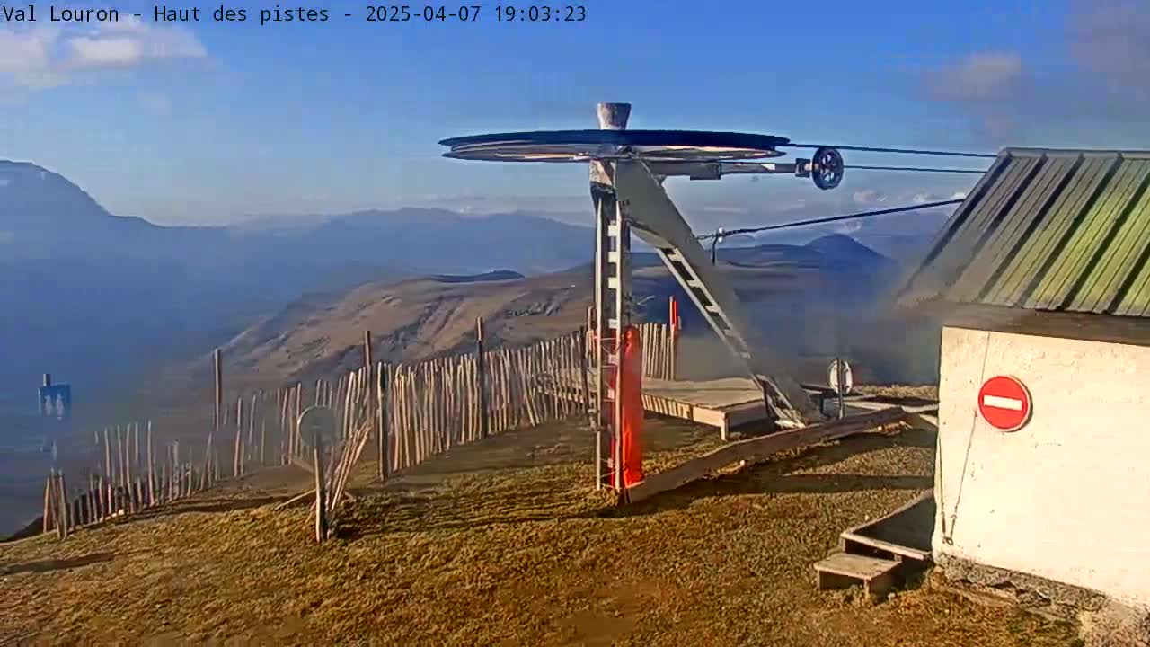 A ski lift pulley system and small building are situated on a mountaintop with a clear, sunny sky and distant hazy mountains visible.