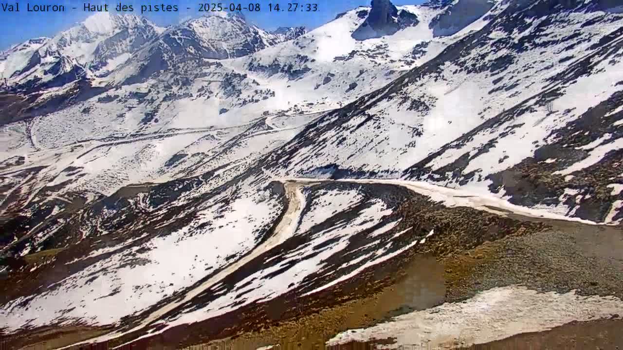 A winding mountain road is partially covered in snow under a sunny sky.