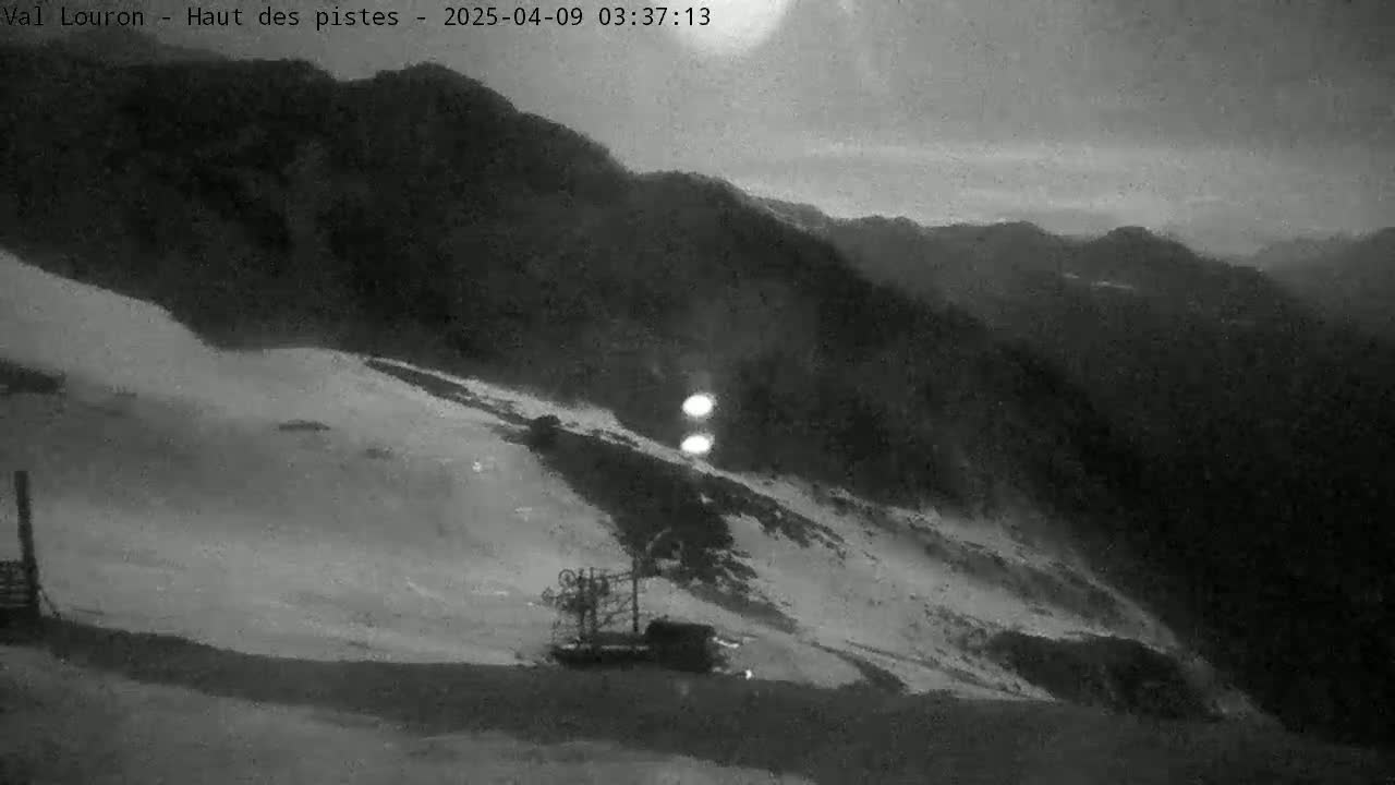 A nighttime, grayscale view shows a snow-covered slope with a ski lift and mountains in the background under a dark, cloudy sky.