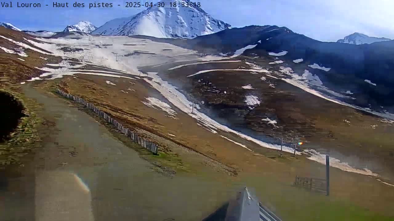A mountain road winds through a mostly snow-free, brown and green hillside, with patches of snow remaining on the upper slopes under a clear blue sky.