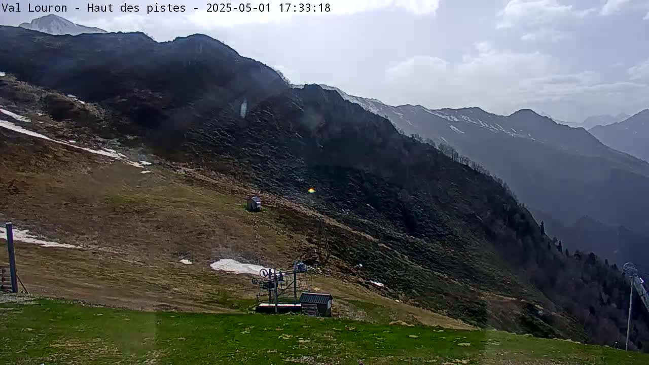 A mostly clear day shows a mountainside with patchy snow, a ski lift, and small buildings; mountains stretch into the distance under a partly cloudy sky.