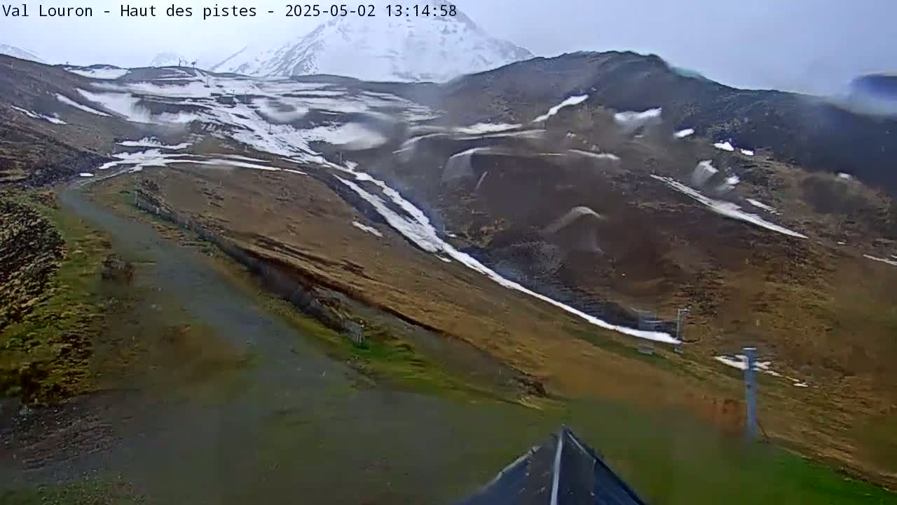 A mountainside with patches of snow and brown grass is seen on an overcast day, with some snow blowing in the wind.