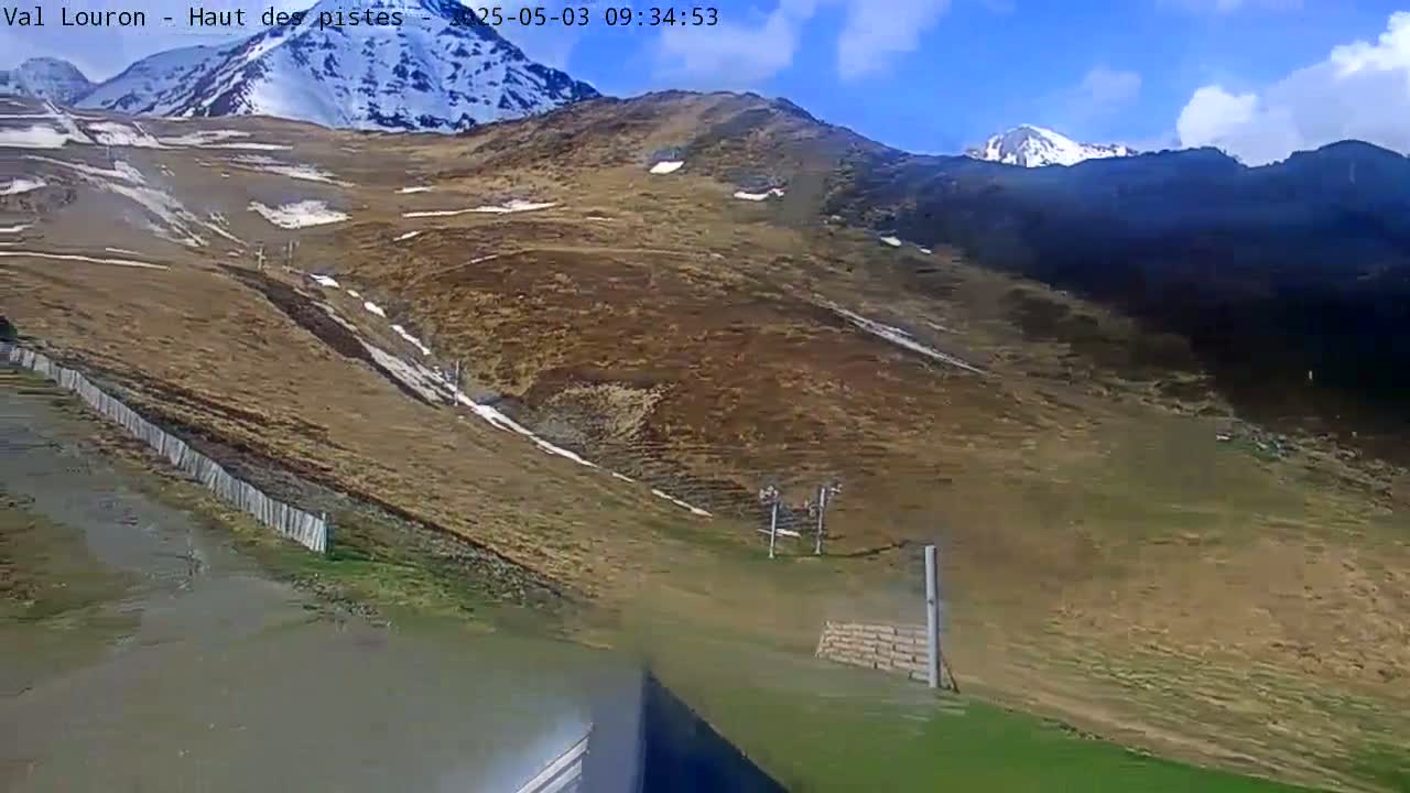 A mostly brown and tan mountainside with patches of snow and a partly cloudy blue sky is shown, with a few ski lifts visible.