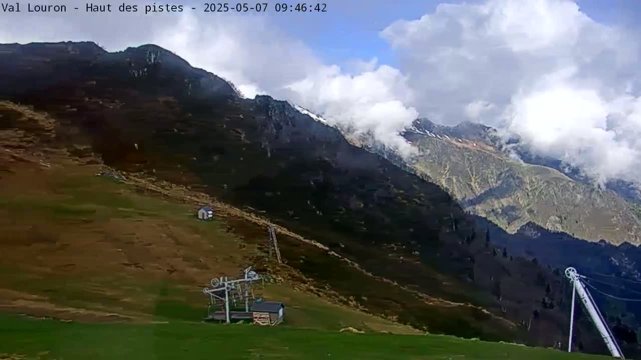 A partly cloudy day reveals a mountainous landscape with a ski lift and a small building situated on a grassy slope below the peak.
