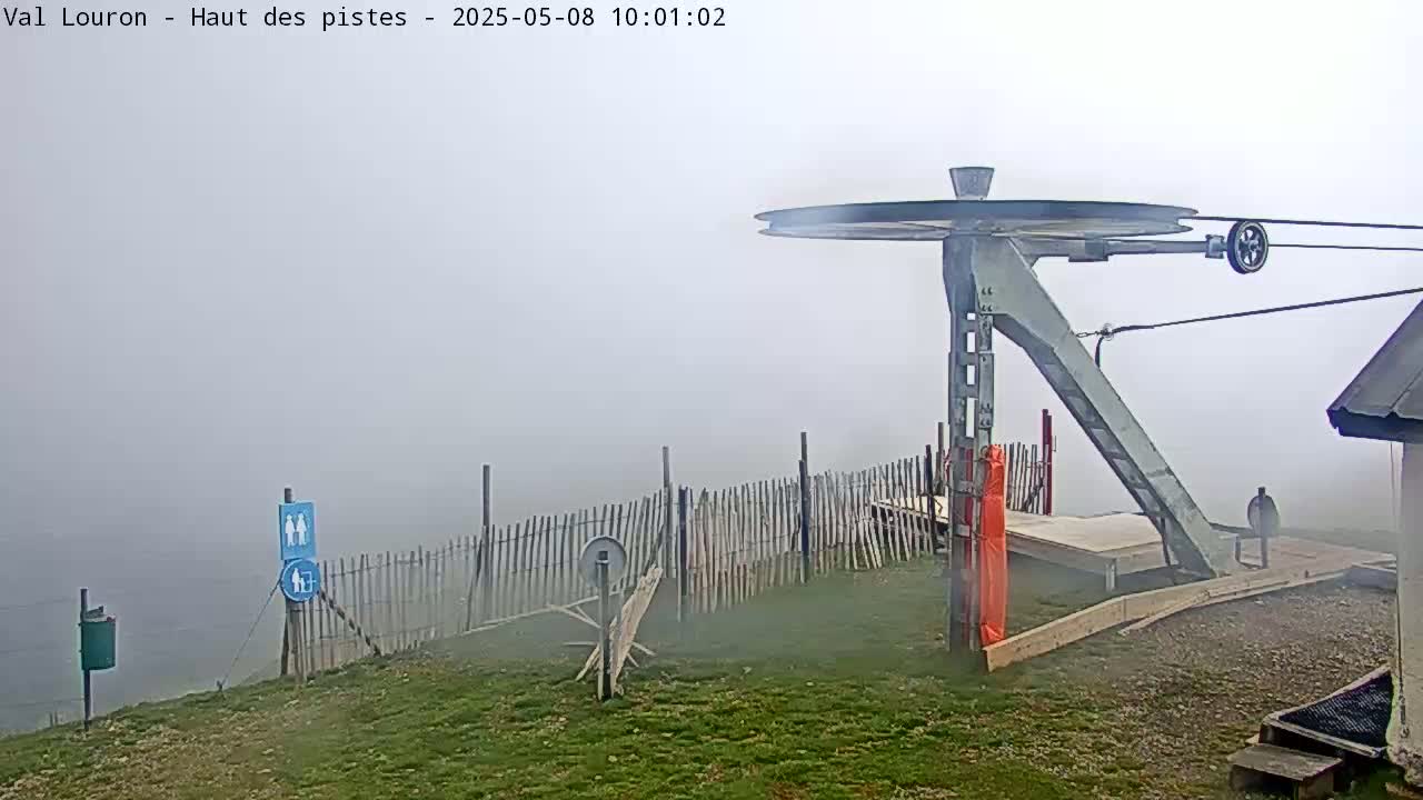 A ski lift top terminal is shrouded in fog, with a wooden fence and grassy area visible in the foreground.
