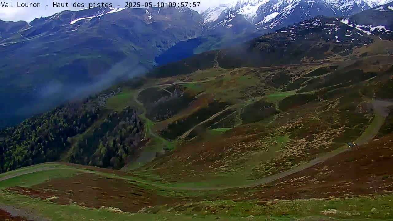 A high-angle view shows a mountainous landscape with winding dirt roads, patches of green and brown vegetation, and snow-capped peaks in the background under hazy conditions.