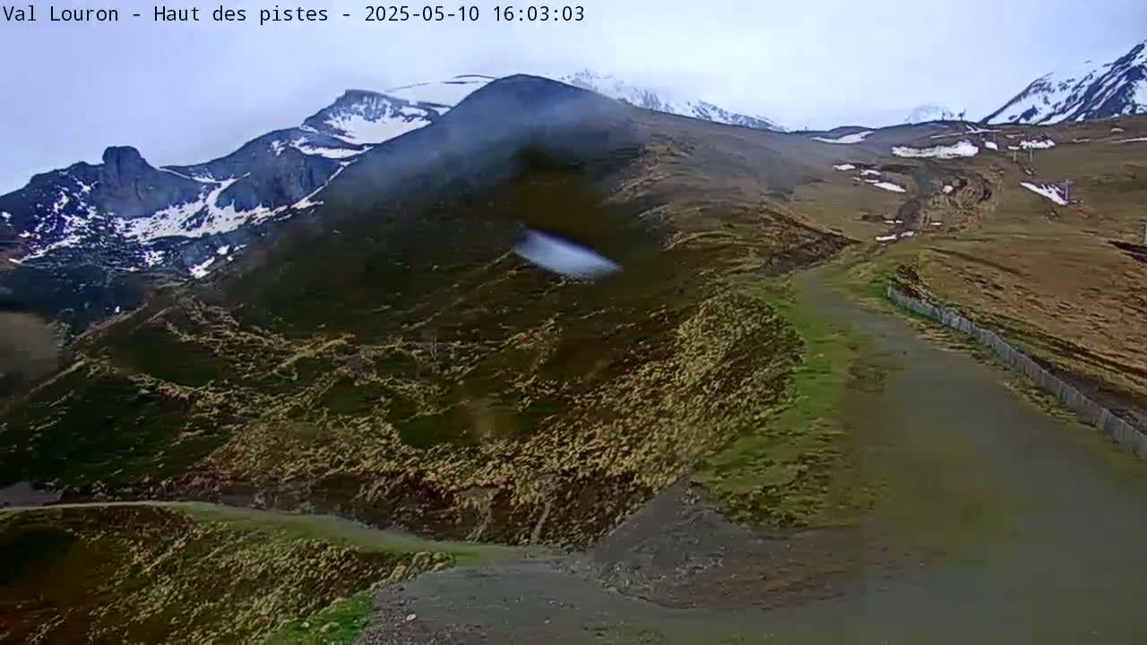 A winding mountain road leads upward toward snow-capped peaks under an overcast sky.