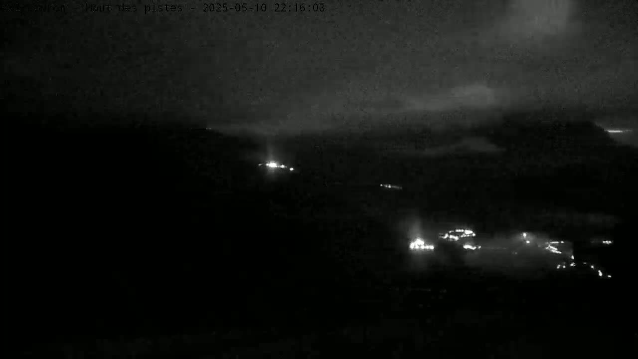 A nighttime, low-light view shows a distant landscape with a few scattered points of light, possibly buildings or vehicles, under a cloudy sky.