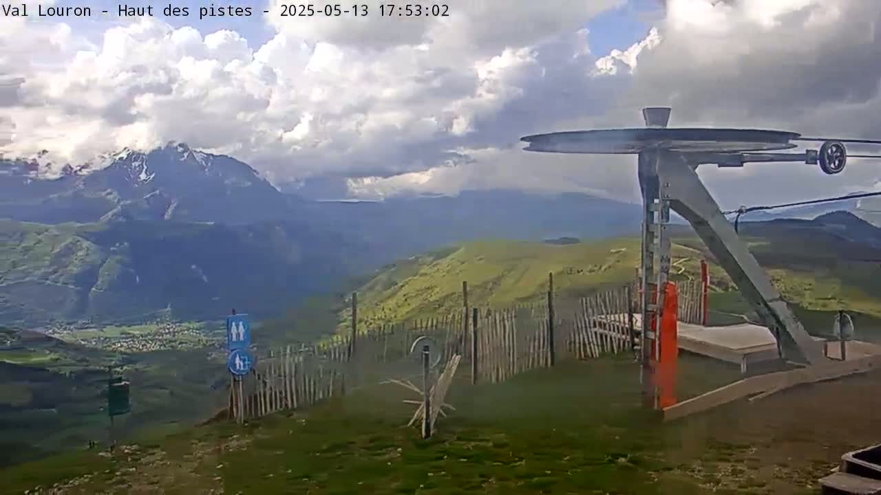 A partly cloudy sky overlooks a mountaintop view of a valley and snow-capped mountains, with a ski lift pulley system in the foreground.
