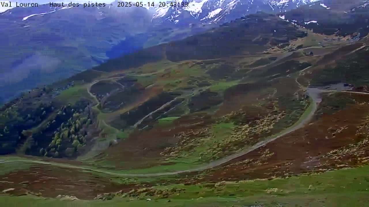 A winding road curves through a mountainous landscape of green and reddish-brown hills, with snow-capped peaks visible in the distance under hazy skies.
